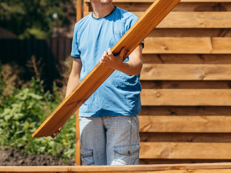 A homeowner or contractor holding a wooden fence board during a backyard fence building project.