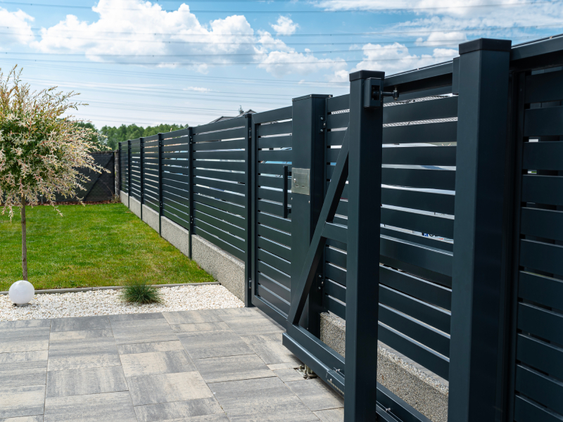 Modern dark grey sliding driveway gate with wide horizontal slat panels and an integrated pedestrian gate set along a concrete curb and paved stone driveway.