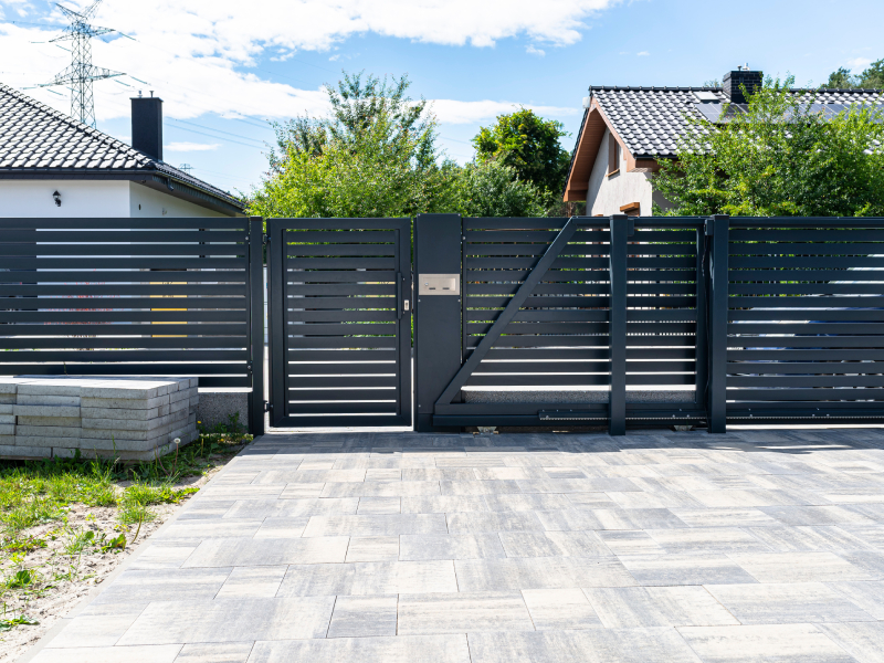 Modern dark grey metal fence featuring horizontal slats, a matching walkthrough pedestrian gate, and a wide sliding driveway gate set along a paved stone driveway.